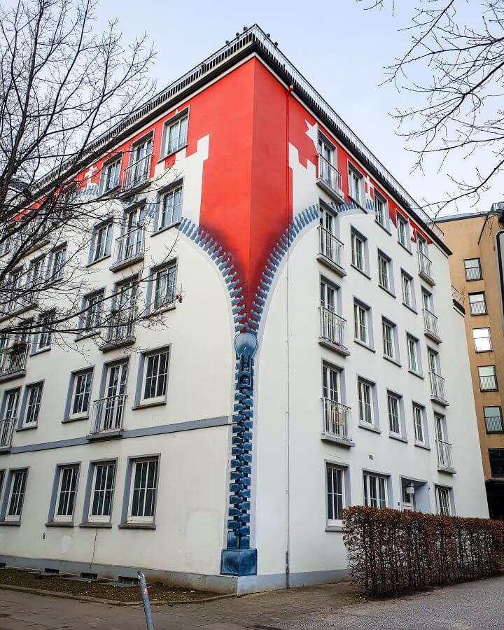 A building on the streets of Hamburg, Germany that is painted to look like the white is being zipped off to see a red building underneath "How to See Hamburg on a Budget"