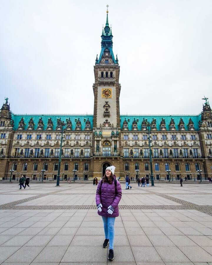 Krissie in a big purple coat standing in front of the impressive Hamburg town hall with its glorious green roof "How to See Hamburg on a Budget"