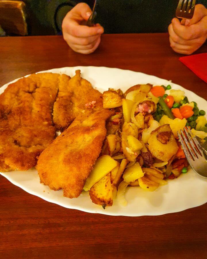 An impressive looking schnitzel, potatoes cooked with onion and bacon and some veggies on a large plate. In the background Krissie is seen holding a knife and fork ready to eat it all! "How to See Hamburg on a Budget"