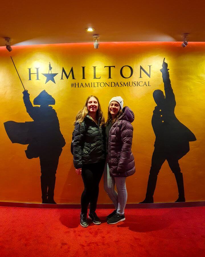 Katherine and Krissie with big smiles standing in front of a sign that says Hamilton #HamiltonDasMusical in Hamburg, Germany "How to See Hamburg on a Budget"