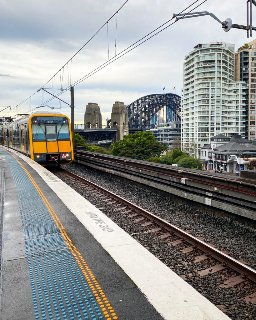 Four Easy Day Trips From Sydney on the Train. A train coming in to the platform with a yellow front and a blue and yellow line on the platform letting people know how far back to stand. Behind the train is a view of the Sydney Harbour Bridge, some trees and some apartment blocks.