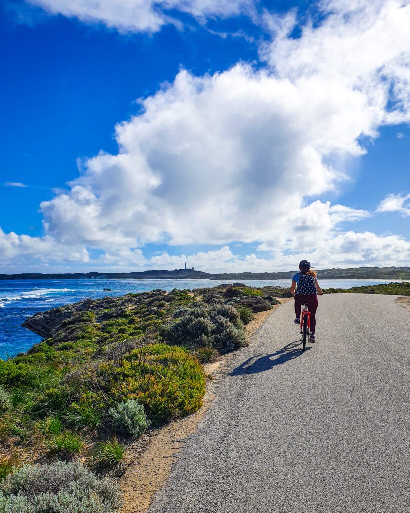 Krissie on the right of the photo riding on a bike down a grey path on Rottnest Island, WA. Beside her on the left is some low green bushes and the crystal blue water. Beyond her is more island with a lighthouse in the distance "Rottnest Island Day Trip: Everything You Need to Know"