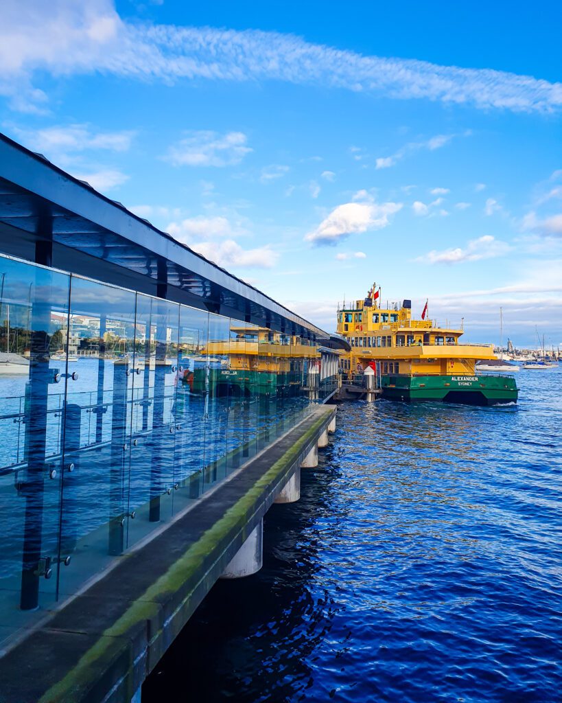 A glass walkway along the left of the photo and water on the right with a ferry docking at the end in Sydney. The ferry is yellow on top and green on the bottom and is 3 levels high "A Complete Guide to the Public Transport in Sydney"