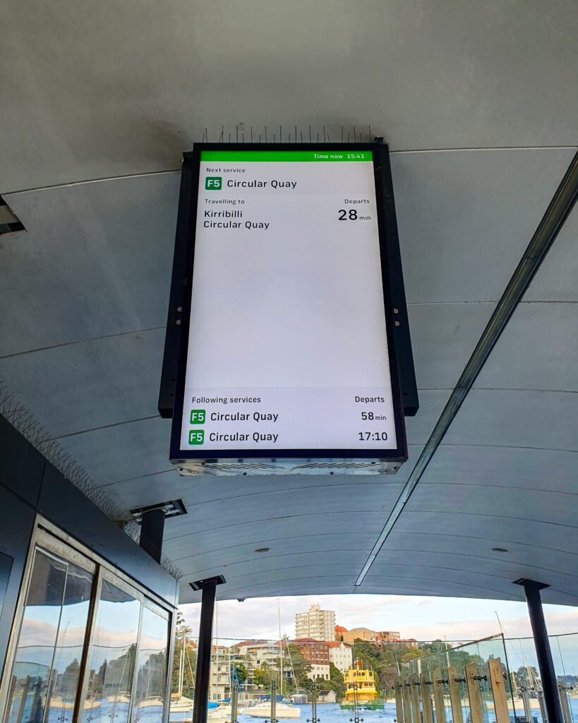 A big electronic board stuck on the roof of the ferry terminal that says the ferry is first going to Kirribilli and then Circular Quay in Sydney. Under the board is some glass walls and you can see a bit of the harbour with a ferry in the distance "A Complete Guide to the Public Transport in Sydney"