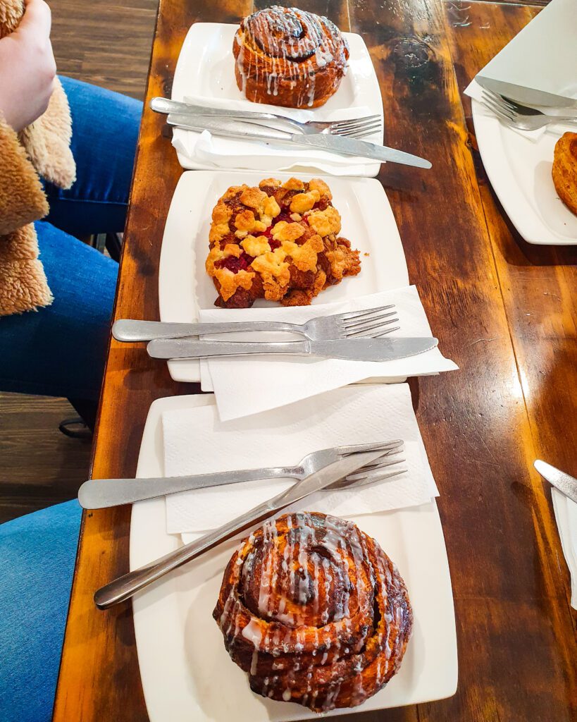 A brown wooden table with two round crispy cinnamon scrolls with white icing on top on white plates. On a plate in between is a croissant with cookie bits in it and all the plates have knifes and forks on them and they were from the Old Cookie Factory in Adelaide, SA "How to See the Best of Adelaide in a Weekend"