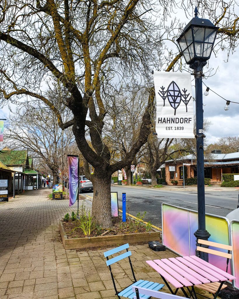 A street in Hahndorf, South Australia with a lamppost and a sign on it that says "Hahndorf EST. 1839". Below the signpost is a colourful table and some chairs and behind it are trees, shops and cars "How to See the Best of Adelaide in a Weekend"