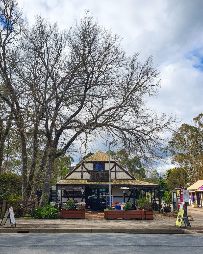 A little white building with wooden beams in it and a pointed roof in Hahndorf, South Australia. The photo is taken from across the road and beside the building are some trees "How to See the Best of Adelaide in a Weekend"