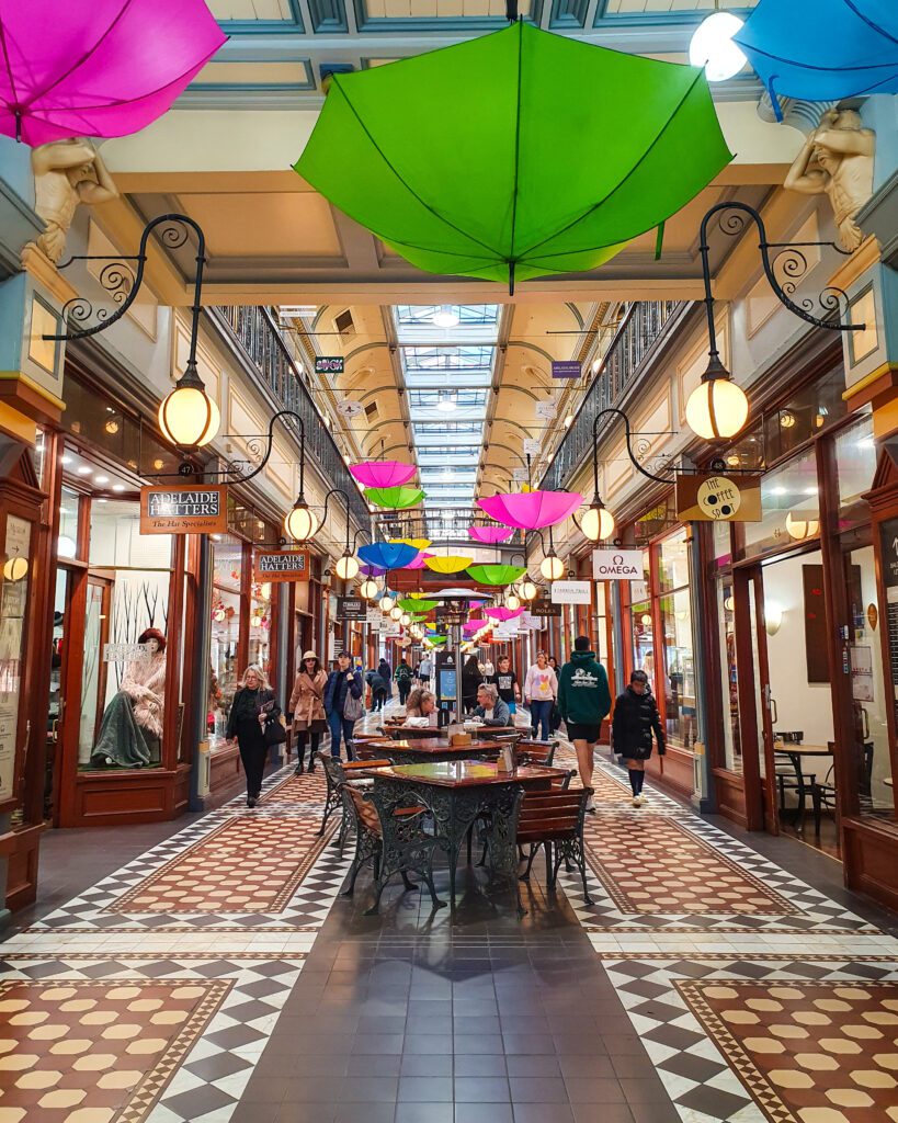 Inside the beautiful Regent Arcade in Adelaide, SA with old style brown, white and black tiling on the floor and brown exteriors to all the shops on either side of the arcade. Above the shops are some coloured umbrellas hanging from the roof "How to See the Best of Adelaide in a Weekend"