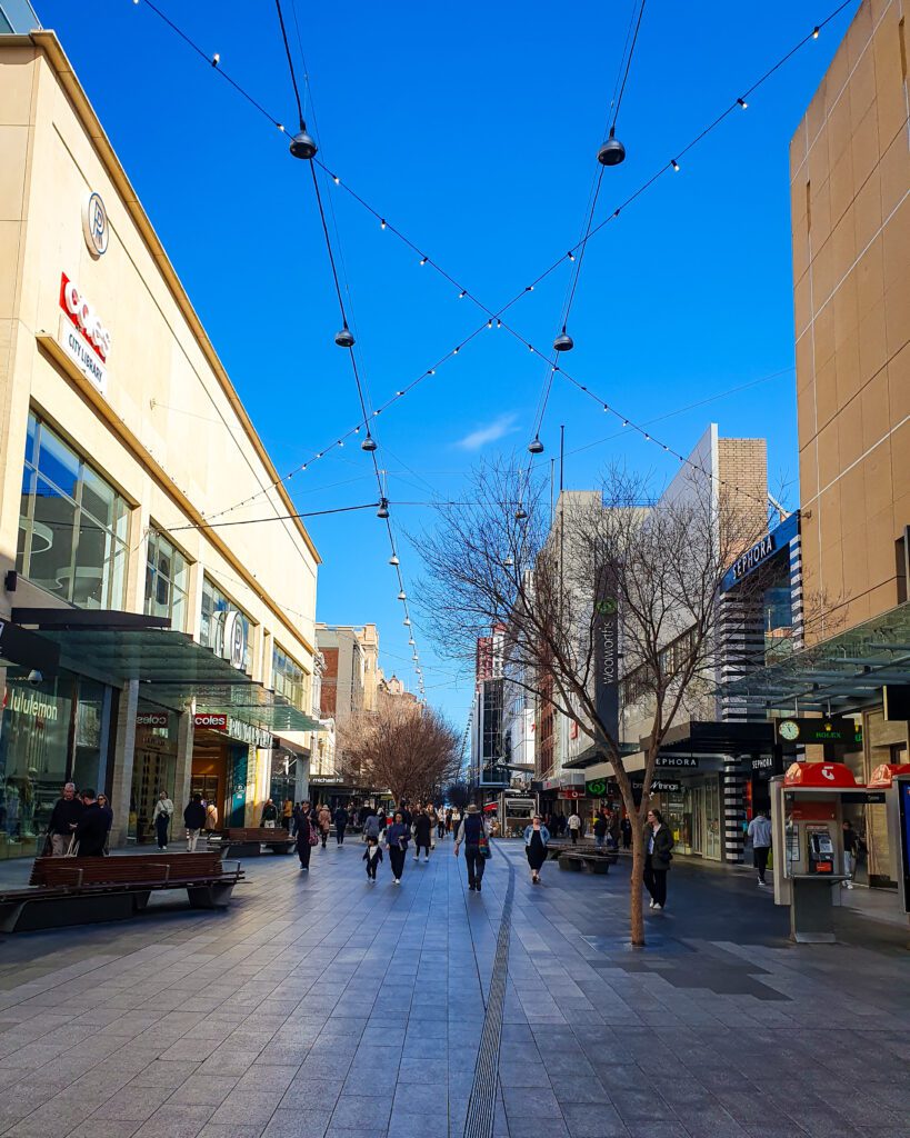 Rundle Mall in Adelaide, SA that is a long outdoor walkway with shops on either side. Above the path are some lights strung between the shops and above it all is a beautiful blue sky "How to See the Best of Adelaide in a Weekend"