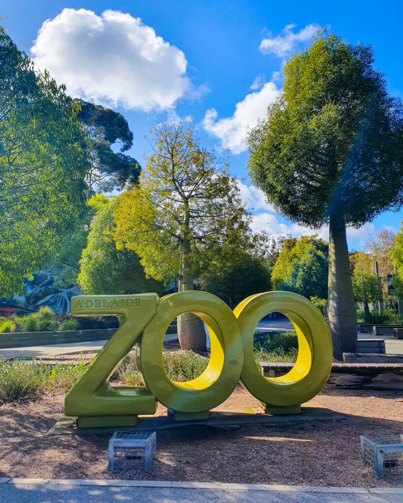 A giant gold sign outside Adelaide Zoo, SA that says ZOO with Adelaide etched into the Z of the sign. Behind the sign are some trees and the blue sky "How to See the Best of Adelaide in a Weekend"