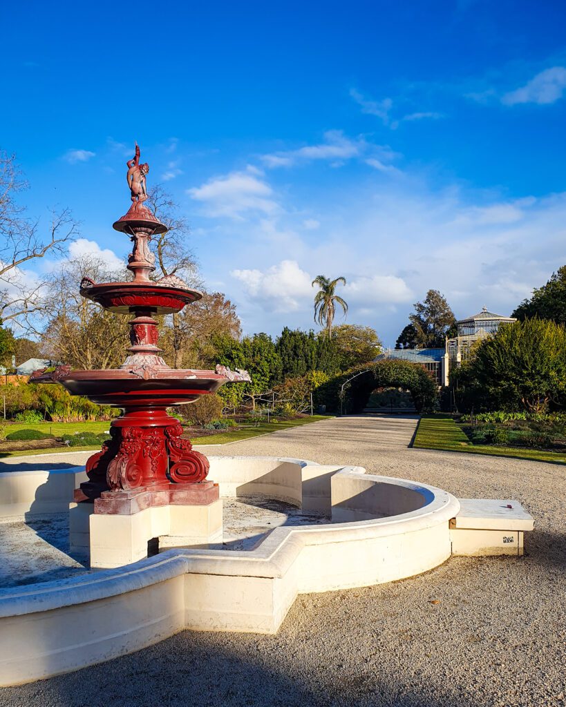 Inside the Adelaide Botanic Gardens, SA with a white stone pond that has a red fountain in the middle of it. Behind the fountain are lots of green plants and the top of a glass greenhouse can be seen "How to See the Best of Adelaide in a Weekend"