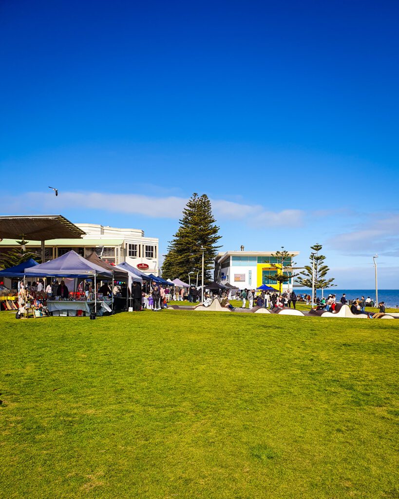 Some tents with market stalls on a big patch of green grass next to Henley Beach, Adelaide SA. On the right you can see the ocean and behind the stalls are some buildings and trees "How to See the Best of Adelaide in a Weekend"