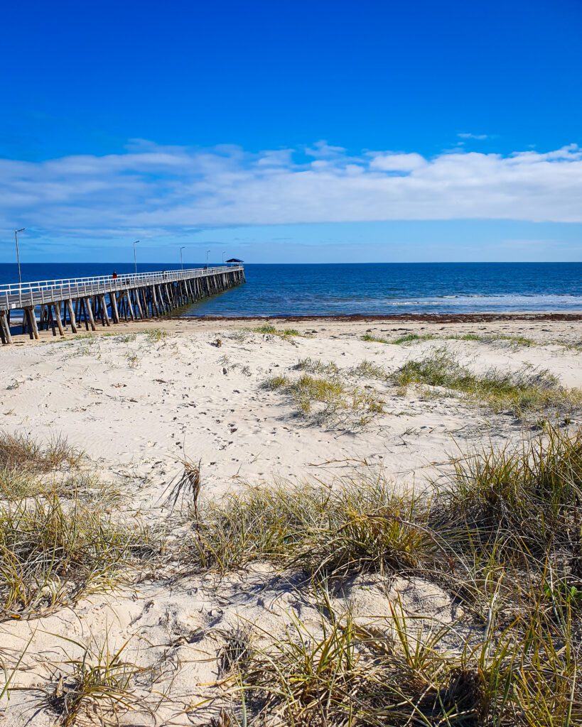 The bottom half of the photo is sand and grass on Grange Beach, Adelaide SA. Beyond that is water and the blue sky and on the left of the photo is a long wooden jetty that goes out to sea "How to See the Best of Adelaide in a Weekend"