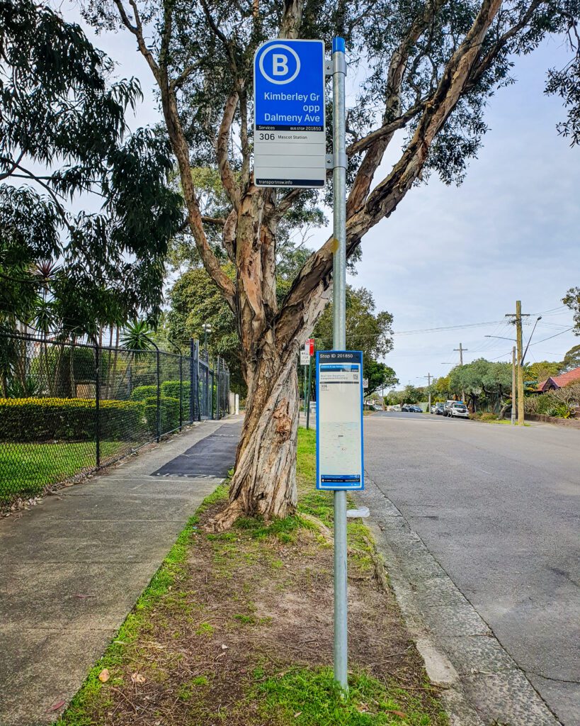 A suburban street with trees and grass in between the path and road with a metal pole with two signs on it. The bottom sign is unreadable but the top one has a big B in a circle and the name of the street on it in Sydney "A Complete Guide to the Public Transport in Sydney"