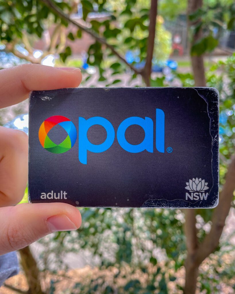 A hand holding a black Opal card that says Adult and NSW down the bottom. Behind it is a blurred background of trees "A Complete Guide to the Public Transport in Sydney"