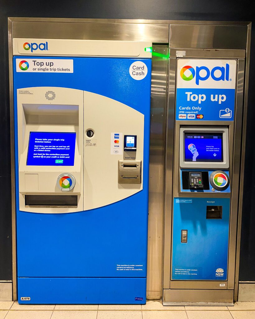 A big silver and blue machine in Sydney with a screen to purchase an Opal card. Next to it is a smaller screen with a sign above that says "Opal Top Up Cards Only" "A Complete Guide to the Public Transport in Sydney"