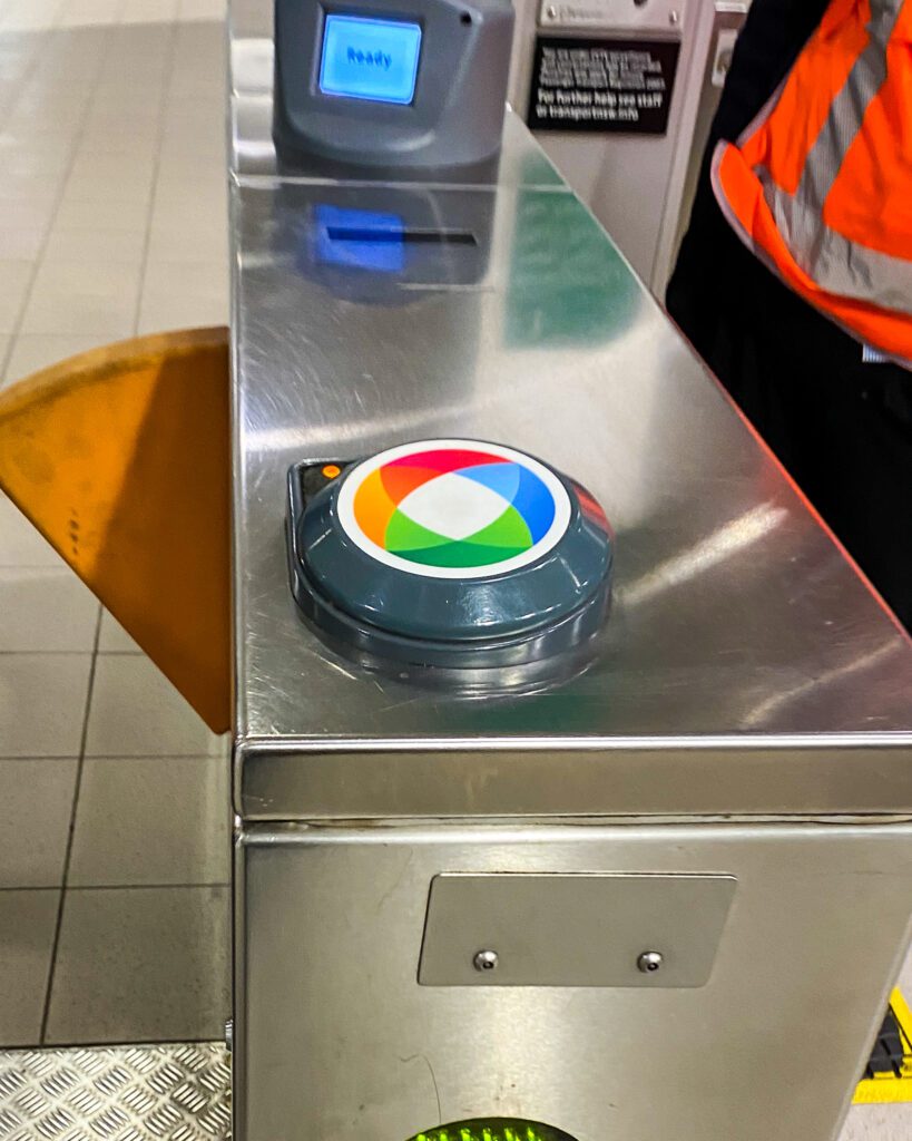 A ticket gate at a train station in Sydney with a orange, red, green and blue circle on a big silver barrier next to the gate where you tap your Opal card to get through "A Complete Guide to the Public Transport in Sydney"