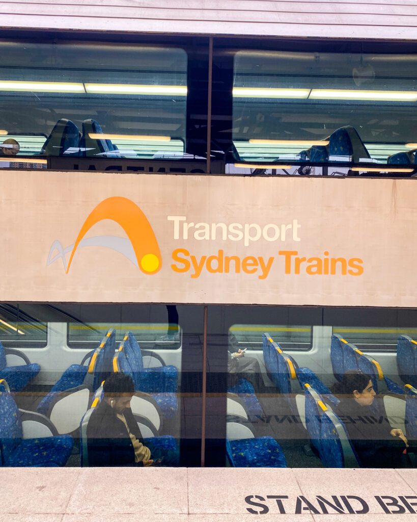 A Complete Guide to the Public Transport in Sydney. A photo of a portion of a Sydney train with people sitting in it. You can see a corridor upstairs with chairs and one downstairs and in the middle is a bar that says "Transport Sydney Trains"