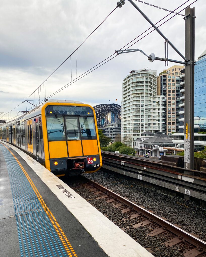 A train coming in to the platform with a yellow front and a blue and yellow line on the platform letting people know how far back to stand. Behind the train is a view of the Sydney Harbour Bridge, some trees and some apartment blocks "A Complete Guide to the Public Transport in Sydney"