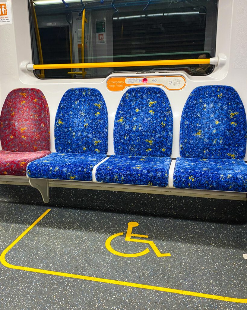 4 chairs along the wall on a Sydney train. 3 of the chairs are blue with a yellow box on the floor with a yellow disabled sign in the middle and the other chair is red "A Complete Guide to the Public Transport in Sydney"