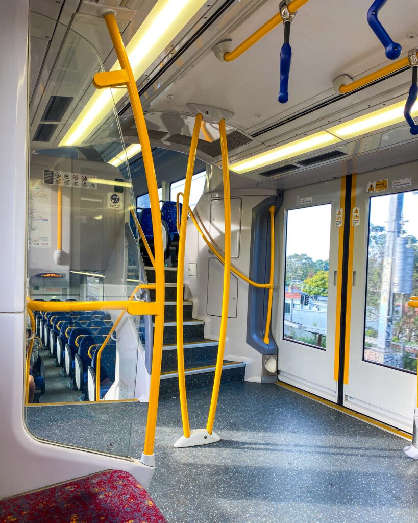 A photo taken inside a Sydney train on the middle level. You can see stairs leading up to a top level and stairs leading down to a bottom level with lots of yellow bars everywhere to hold onto "A Complete Guide to the Public Transport in Sydney"