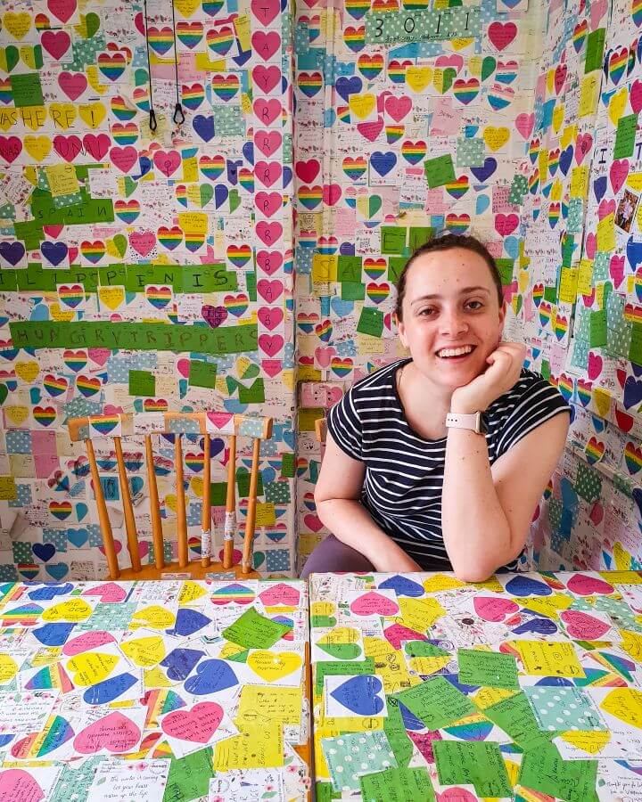 Krissie sitting at a table in the Note Coffee shop and smiling at the camera in Hanoi, Vietnam. Next to her is a spare wooden chair and the rest of the space around her including on the table and all over the walls is taken up by colourful post it notes "Explore Hanoi: The Most Wonderful Budget Destination"