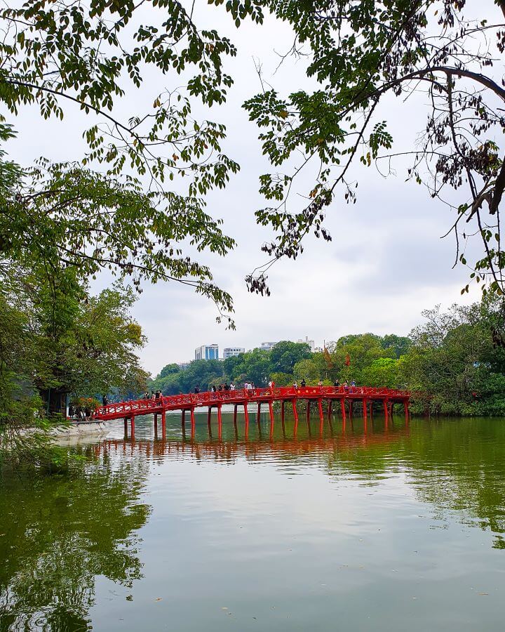 A lake in Hanoi, Vietnam with a red foot bridge going from one side to an island in the middle. On both sides are lots of green trees including in the foreground and in the background some high rise buildings can be seen "Explore Hanoi: The Most Wonderful Budget Destination"