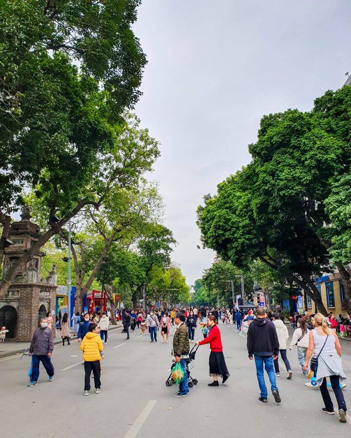 A street in Hanoi, Vietnam with big green trees on either side and lots of people walking on the road. This was taken during the walking only time for some of the main roads in Hanoi "Explore Hanoi: The Most Wonderful Budget Destination"