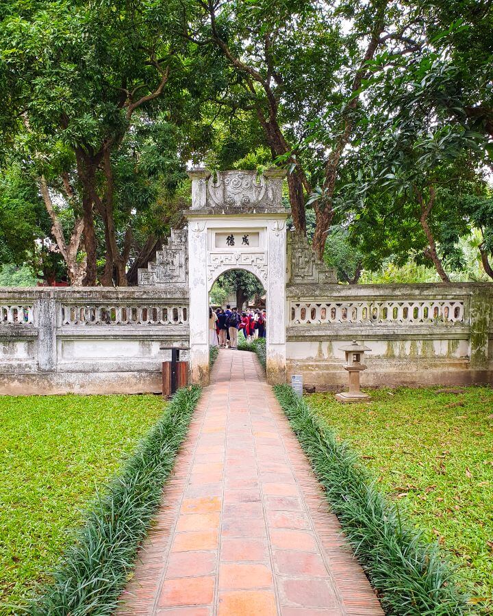 A brick walkway in the Temple of Literature in Hanoi, Vietnam. On the side of the pathway is neat grass and at the end is a stone wall and archway. On the other side of the wall are lots of green trees and some people "Explore Hanoi: The Most Wonderful Budget Destination"