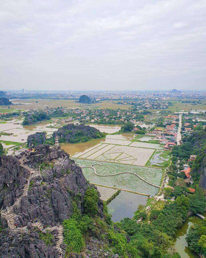 A view of the Ninh Binh Region in Vietnam over the rice paddies that are full of water and another view point lower down on the mountain with some stairs and people on it. In between the paddies are little limestone mountains, lots of green plants and lots of little buildings with orange roofs "Explore Hanoi: The Most Wonderful Budget Destination"