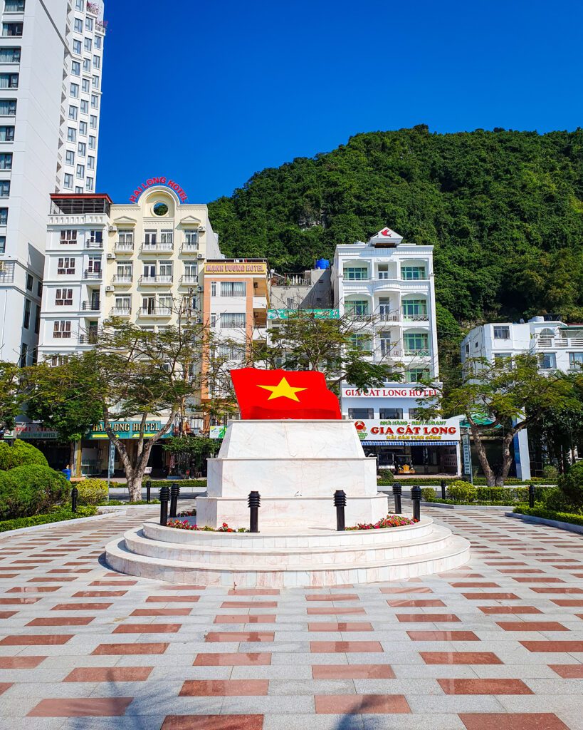 A town square on Cat Ba Island in Vietnam. The square has red and white tiles on the floor and a statue in the middle of the Vietnam flag on a plinth. Behind the square are some thin multi level buildings and a green mountain behind them "Lan Ha Bay: A Beautiful Alternative to Halong Bay"