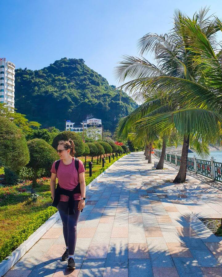 Krissie walking on a tiled path with a fence, some palm trees and the water on the right of the photo on Cat Ba Island, Vietnam. On the left is lots of lush green trees and grass and behind her is a big green hill and some buildings "Lan Ha Bay: A Beautiful Alternative to Halong Bay"