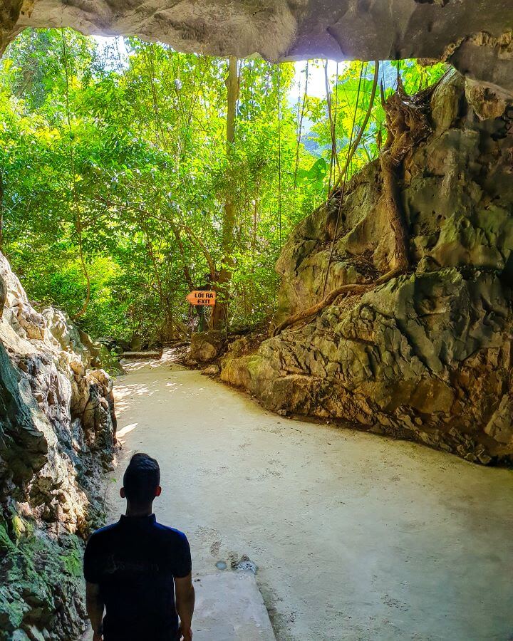 The silhouette of our guide showing us out of a cave on Cat Ba Island in Vietnam. We're in the mouth of the cave so there is rock on all sides and the green forest at the end of the mouth with a sign saying EXIT "Lan Ha Bay: A Beautiful Alternative to Halong Bay" 