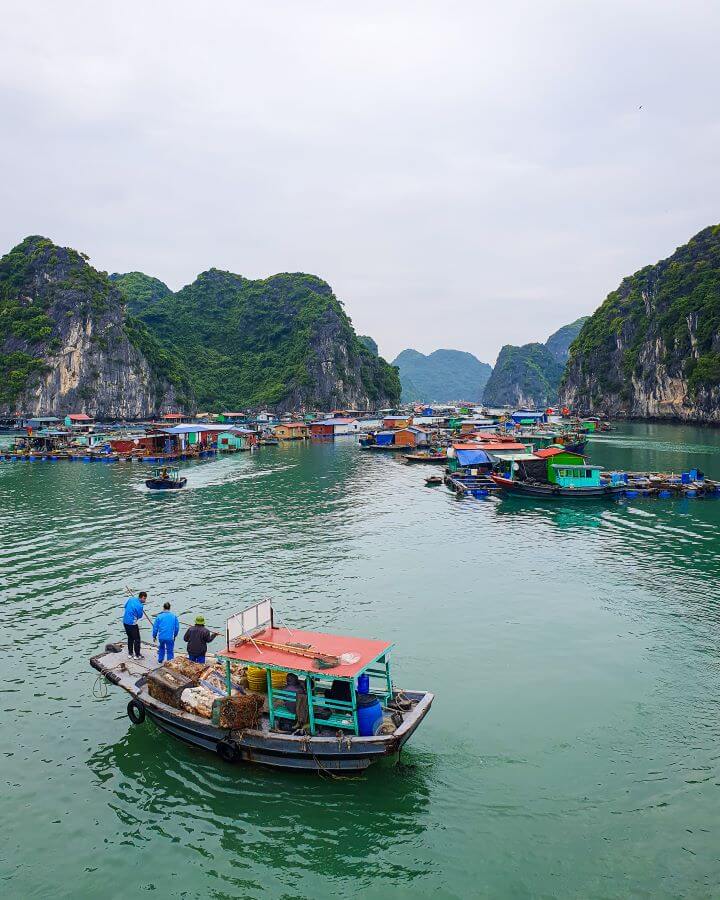 The blue green water of Lan Ha Bay in Vietnam with limestone hills covered in green bushes in the distance. In the foreground is a little wooden boat with a red roof and people standing in it, and behind that is a large floating village. The little houses are all different colours and there are boats and people dotted around "Lan Ha Bay: A Beautiful Alternative to Halong Bay"