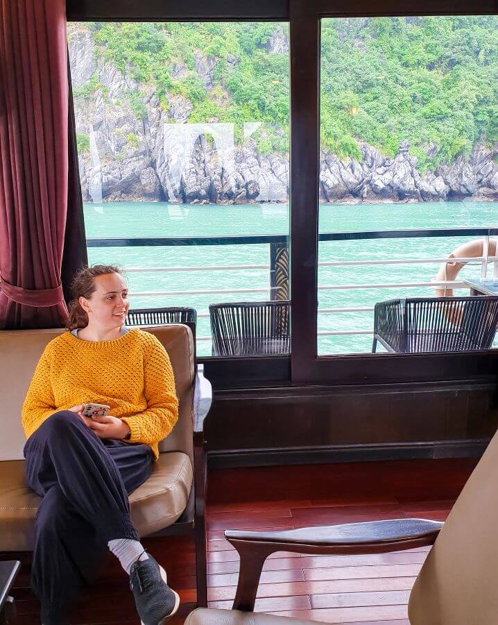 Krissie sitting on a comfy looking couch inside the boat on Lan Ha Bay in Vietnam. Next to her is a another chair and behind her is a red curtain and a big window showing some outdoor chairs and the railing of the boat, with the water and limestone hills of Lan Ha Bay behind them "Lan Ha Bay: A Beautiful Alternative to Halong Bay"