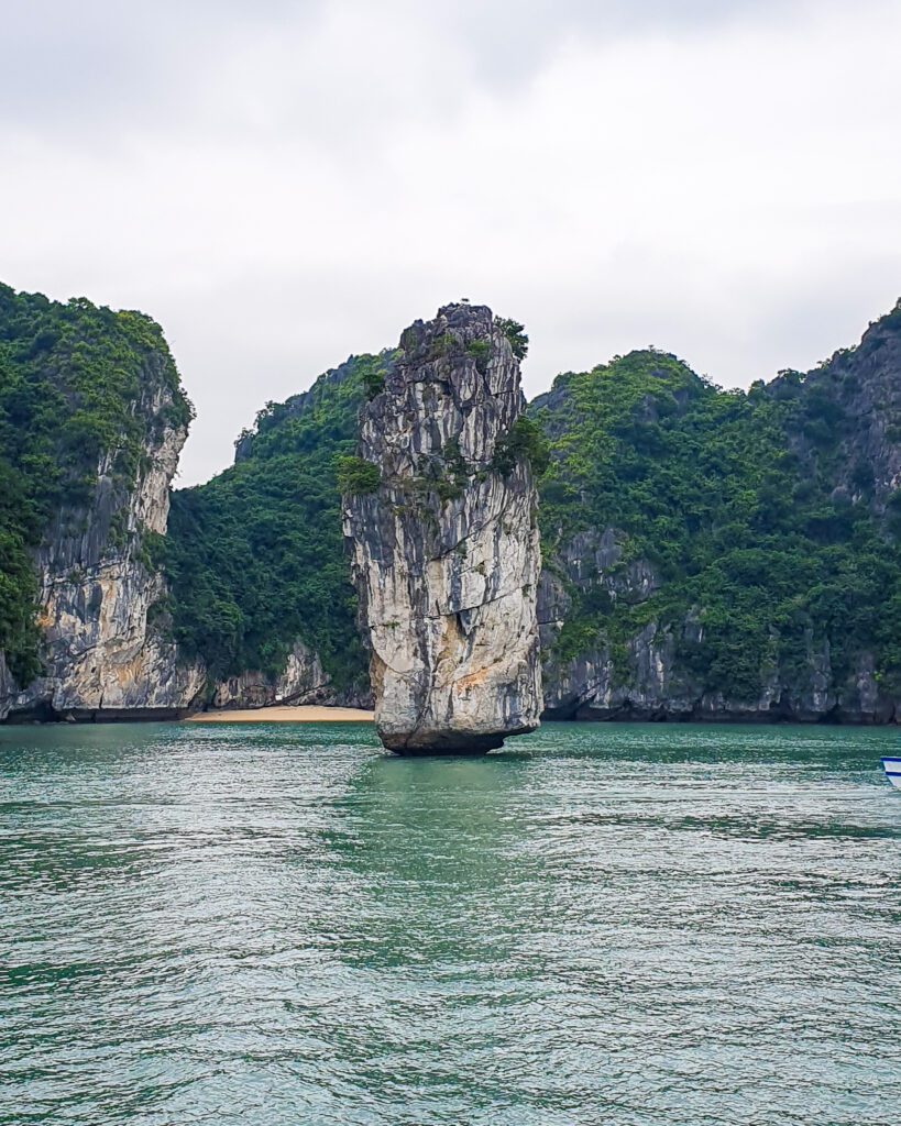 A limestone sliver in Lan Ha Bay, Vietnam, that's been worn away by years of water battering it. It's standing tall with only water around it and the bottom has been worn away so that it gets thicker the higher up it goes. Behind it are some more limestone hills with green bushes and a little beach leading to the water "Lan Ha Bay: A Beautiful Alternative to Halong Bay"