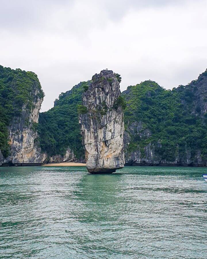 A limestone sliver in Lan Ha Bay, Vietnam, that's been worn away by years of water battering it. It's standing tall with only water around it and the bottom has been worn away so that it gets thicker the higher up it goes. Behind it are some more limestone hills with green bushes and a little beach leading to the water "Lan Ha Bay: A Beautiful Alternative to Halong Bay"