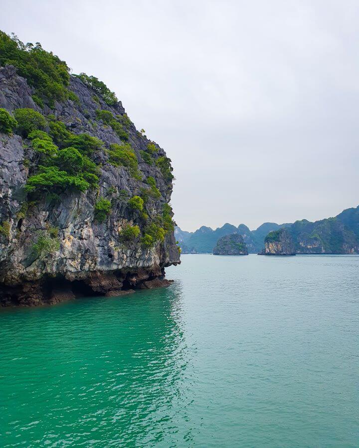 Lan Ha Bay: A Beautiful Alternative to Halong Bay. A picture in Lan Ha Bay, Vietnam, with blue green water and lots of limestone hills in the water with green bushes all over them. The air is kind of hazy and the sky is grey.