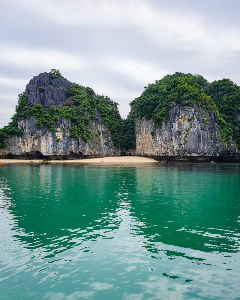 The blue green water of Lan Ha Bay in Vietnam with two limestone hills with green bushes over them. In between the hills is a little beach with yellow sand and it's completely empty of people "Lan Ha Bay: A Beautiful Alternative to Halong Bay"