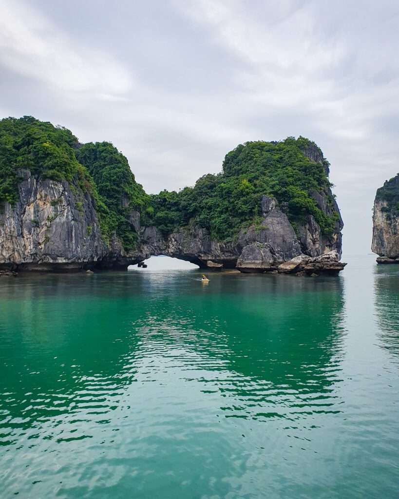 The blue green water of Lan Ha Bay in Vietnam. In the photo is a big limestone rock with green bushes on top and there's a little cave like hole through the rock on the water level creating a sort of natural bridge and almost splitting the rock into two smaller ones "Lan Ha Bay: A Beautiful Alternative to Halong Bay"