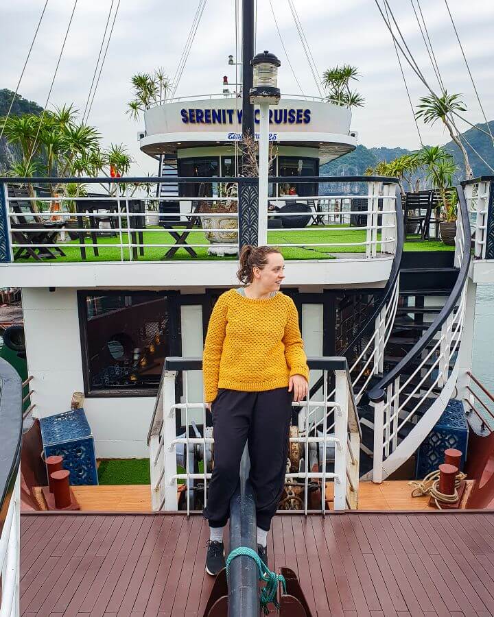 Krissie wearing a yellow jumper and standing on the deck of the boat in Lan Ha Bay in Vietnam. Behind her is the boat with white and black railings, a section on the upper level with fake grass and some green trees and at the top it says "Serenity Cruises" "Lan Ha Bay: A Beautiful Alternative to Halong Bay"