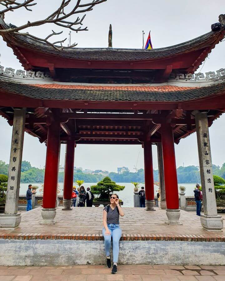 Krissie sitting on the edge of a raised brick platform with a large red and orange pagoda above it at the Ngoc Son Temple in Hanoi, Vietnam. There are potted plants around the platform and in the background the lake can be seen "Explore Hanoi: The Most Wonderful Budget Destination"