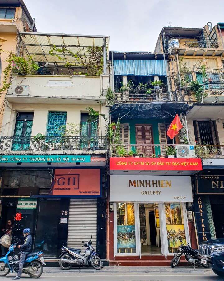 Three thin buildings in Hanoi, Vietnam in different colours with shutters. signs, and plants all over the buildings. The buildings have 3 levels and the bottom level is shops with one of them saying "Minh Hien Gallery". There are also motorbikes out the front of the buildings "Explore Hanoi: The Most Wonderful Budget Destination"