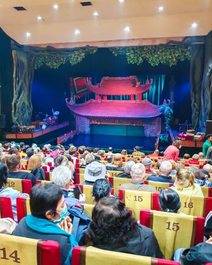 Rows and rows of seats with people sitting in them at the Thang Long Water Puppet Theatre in Hanoi, Vietnam. Behind the seats is a little stage of water with a red pagoda set behind the water and some fake trees and leaves above it. On either side of the water are some platforms with instruments where the musicians and singers will sit "Explore Hanoi: The Most Wonderful Budget Destination"