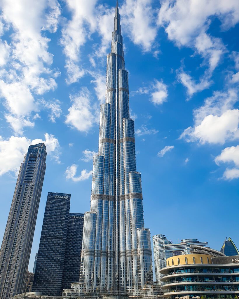 The Burj Khalifa with other buildings around it in Dubai, UAE. The Burj starts out quite thick down the bottom and goes up in tiers until it's a small point in the sky. The other high rise buildings around it come up to about half its height and all around it is a blue, cloudy sky "Nine of the Best Budget Friendly Ways to see Dubai"