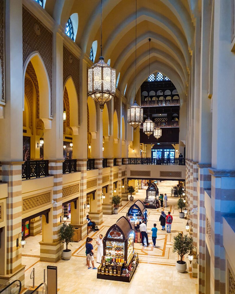 Inside a two level shopping mall in Dubai, UAE, with Arabic style architecture and a high arched sandstone coloured roof. There are people walking around and little stalls on the ground floor selling food and drinks "Nine of the Best Budget Friendly Ways to see Dubai"