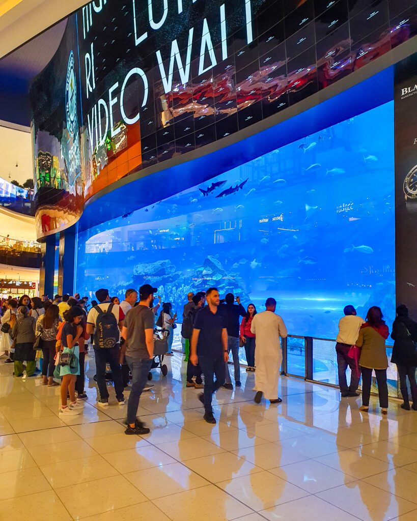 People walking around Dubai Mall, UAE, with a shiny tiled floor. Behind the people is a huge blue water tank with fish and sharks swimming around it "Nine of the Best Budget Friendly Ways to see Dubai"