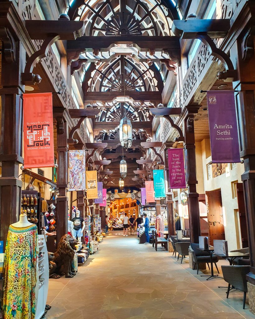 Inside the Souk Madinat Jumeirah shopping area in Dubai, UAE. The area is decorated in an Arabic style with a high arched roof made of dark brown wood. There are shops with clothes and lanterns, chairs and tables and lanterns hanging from the roof too "Nine of the Best Budget Friendly Ways to see Dubai"