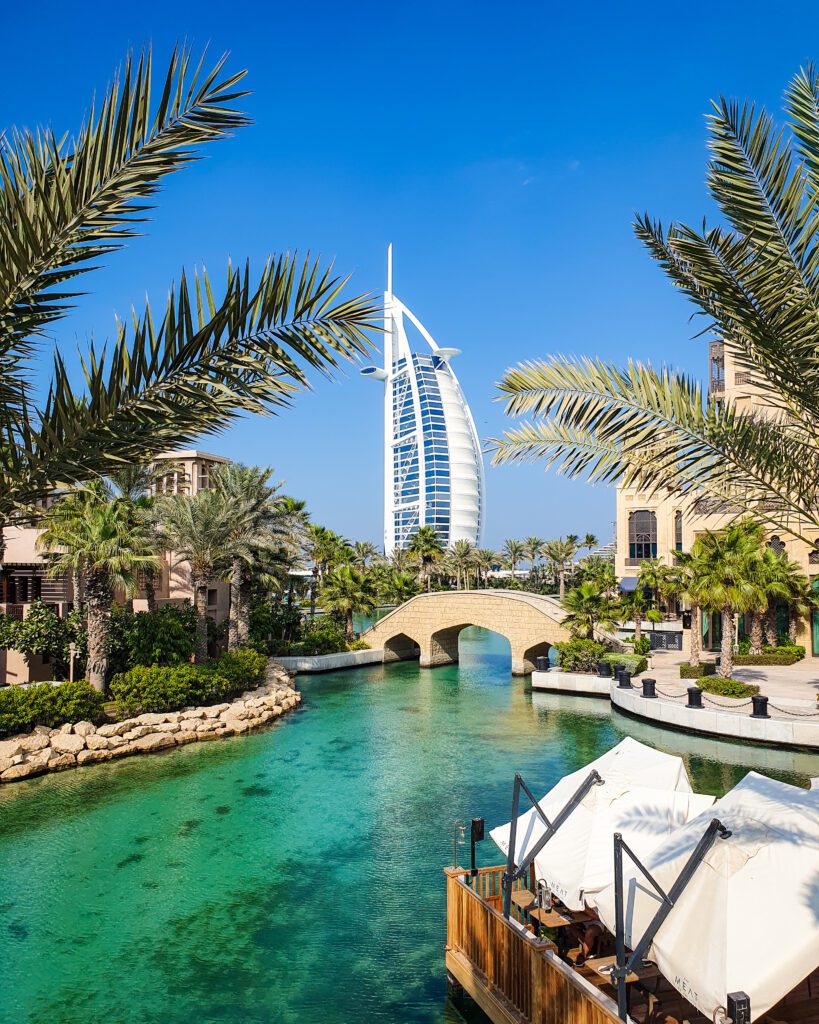 A view of the Burj Al Arab which is a white and blue building in Dubai, UAE that looks like a boat sail. In front of the Burj is a water area with green and blue water, lots of palm trees, a sandstone bridge and a restaurant with big white umbrellas over the tables on the right "Nine of the Best Budget Friendly Ways to see Dubai"