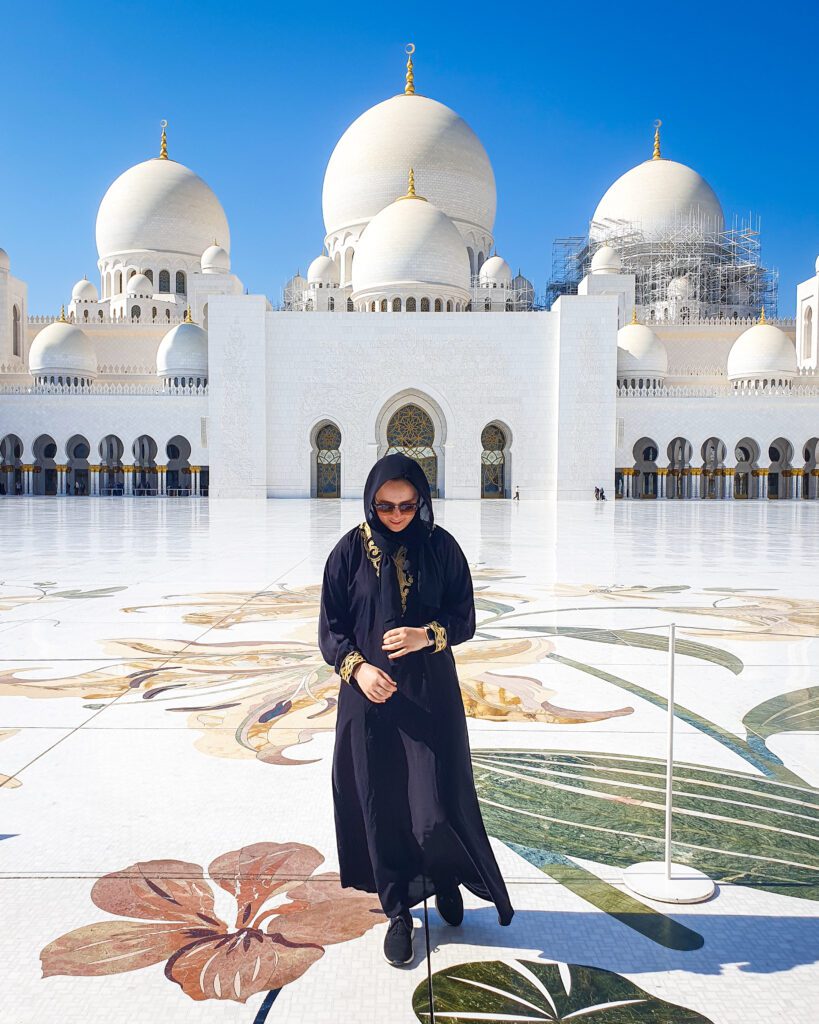 Krissie standing in the large courtyard in the Sheikh Zayed Grand Mosque in Abu Dhabi in the UAE. She's wearing a black Abaya with gold trimming on the wrists and neck that's covering her whole body, leaving only her hands and face bare. She's standing on huge white tiles with green, red and yellow paintings of flowers on them and behind her are three huge white domes with a white building under them and a blue sky above it all "Nine of the Best Budget Friendly Ways to see Dubai"